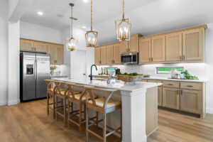 Model home kitchen with stained wood cabinets, quartz island, pendant lighting, and stainless steel appliances.
